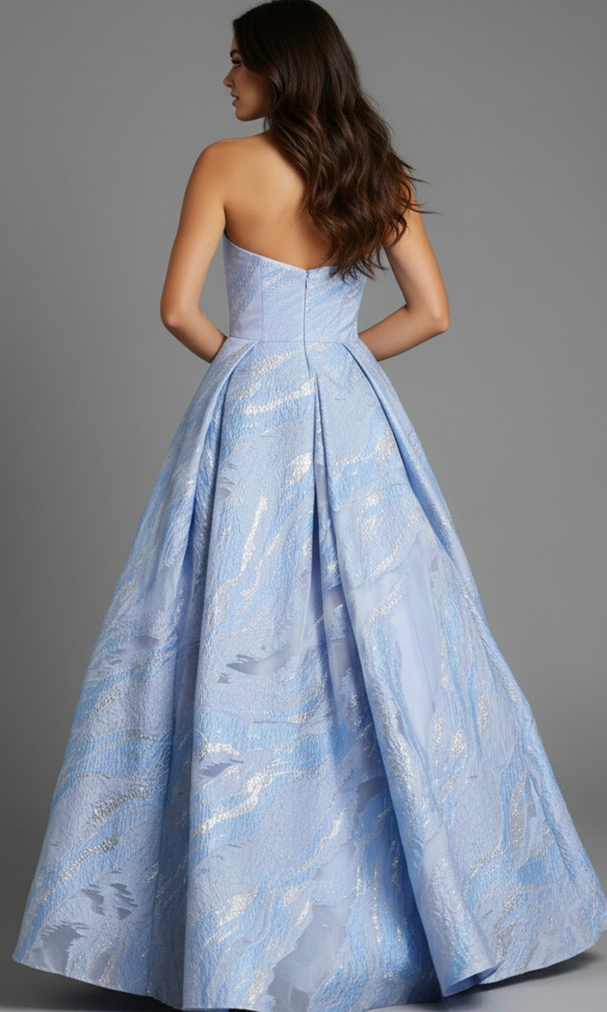 A woman models the Jovani 44369 long prom dress with a strapless, fitted bodice and silver-patterned pleated skirt, standing with her back to highlight the gown’s elegant design.
